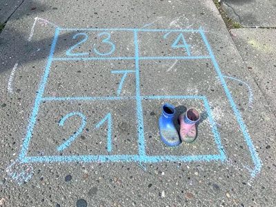 A child's hopscotch grid in blue chalk, numbers wobbly, a pair of small rain boo