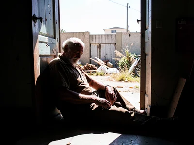 Old man asleep in doorway shadow, brutal noon sun bleaching everything beyond