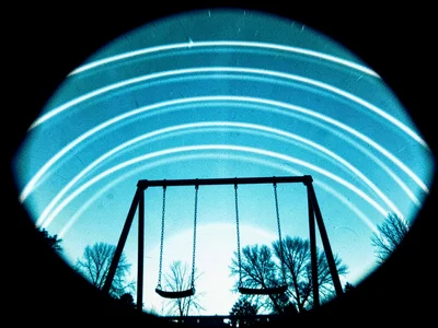 An empty playground swing set, sun arcs crossing in the cyanotype blue sky above
