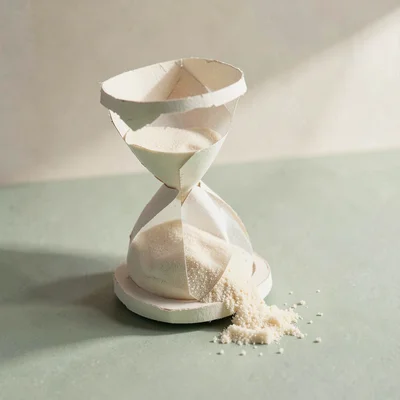 Inside a papercraft hourglass, cream sand spills half out onto a sage tabletop i