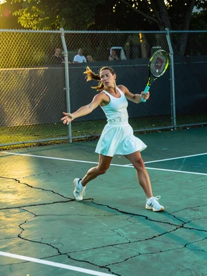 Tennis player mid-serve on cracked court, white dress gone cyan in shade, evenin