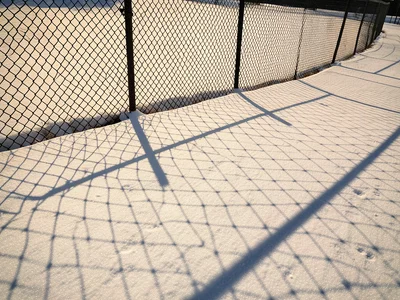 A chain-link fence shadow cast onto fresh snow in low winter afternoon light, th