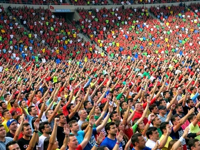 Stadium crowd compressed into bands of color, wave of raised arms rippling throu
