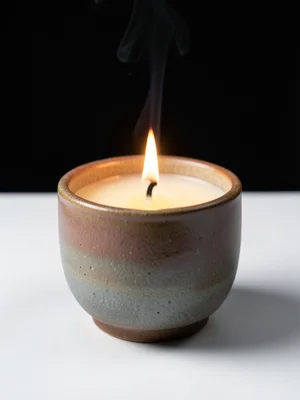 A scented candle in a ceramic vessel, the wax pool and flame visible, a subtle w