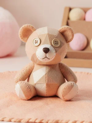 Hand-sewn felt teddy bear with mismatched button eyes sitting upright on a peach