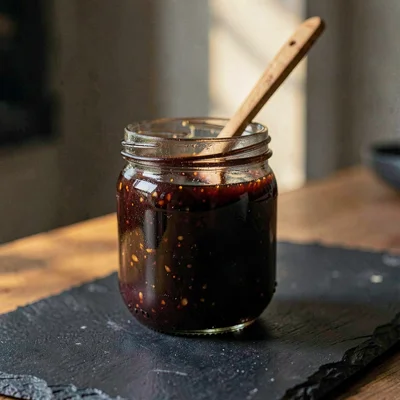 Glass jar of dark fig preserves on dark slate with a wooden spoon perched on top