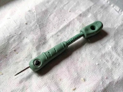 Pricked sage-green sewing needle with a slumped eye and thumb dents along its le