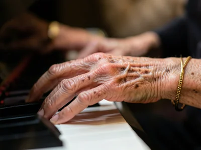 Hands with liver spots and thin skin playing piano, the wrists visible, a gold b