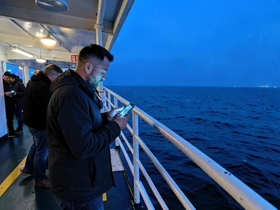 Commuters on ferry deck at blue hour, faces lit by phones against vast blue wate