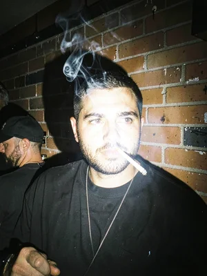 Bouncer's face half-shadowed outside a club at 3am, cigarette smoke frozen mid-e
