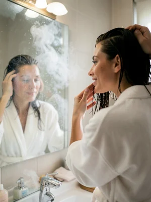 Woman brushing wet hair in front of a steamy bathroom mirror, soft robe, relaxed